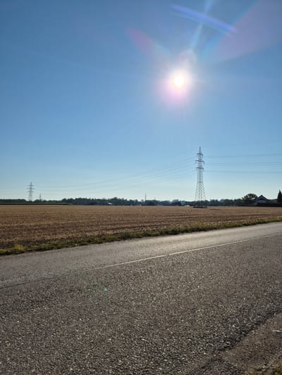 The image depicts a sun-drenched rural landscape with a clear blue sky. In the foreground, a paved road with a white dividing line stretches into the distance. To the left of the road is a harvested field, appearing dry and brown. Several tall, industrial electricity pylons with power lines crisscross the sky over the field and distant tree line. The sun is high in the sky, casting a bright glare and lens flare across the upper portion of the image. In the background, a few houses and trees are visible, suggesting a rural settlement. The scene is peaceful and appears to be taken during daylight hours, likely in the late afternoon due to the angle of the sun and the warm lighting on the road. The location is identified as being in the city of Desselbrunn, Austria. There are no people or animals visible in the image.