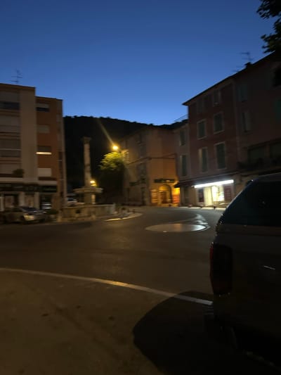 The image depicts a street scene in Anduze, France, during twilight. A tall, multi-tiered stone fountain stands in the center of a roundabout. To the left, a modern apartment building with balconies and shuttered windows is visible. Across the roundabout, a multi-story building with several windows and a shopfront illuminated by interior lights is present. The street is paved, with white and yellow lines marking the road. A vehicle's tailgate and taillight are prominent in the foreground, suggesting the camera is positioned behind a car. The sky is a deep blue, indicating the transition from day to night. The overall atmosphere is quiet and serene, characteristic of a small French town at dusk.