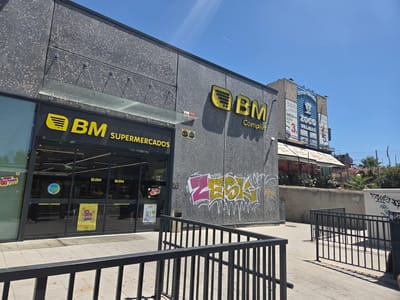 The image shows the exterior of a BM Supermercados store, a supermarket chain in Spain. The store has a modern facade with grey speckled cladding and large glass windows. The BM logo, a yellow stylized leaf shape followed by "BM", is prominently displayed on the building. Below the logo, the word "Complet" is also visible. Inside the store, illuminated signage and shelves stocked with products can be seen through the windows. There are also promotional posters visible, one with "30%" highlighted, indicating a sale or discount.
The store is situated in Collado Villalba, a municipality in the Community of Madrid, Spain. A billboard advertising "ZOCO VILLALBA" and services like "COLADA EXPRESS" (express laundry) is visible in the background, to the right of the BM store, confirming the location. Graffiti art is present on the exterior wall of the BM building, featuring stylized lettering in yellow and pink. In the foreground, black metal railings form a barrier or walkway. The sky is clear and blue, suggesting a sunny day. The overall scene depicts a commercial area on a bright day.