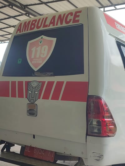 The image shows the rear of a white ambulance parked outdoors under a metal roof structure. The ambulance has "AMBULANCE" written in red letters on its upper rear window. Below this, a shield-shaped emblem with "119" and "Emergency Call ACEH UTARA" is visible. A stylized Transformer logo is also present on the side of the ambulance. The rear lights are red and visible. A red license plate with white text "BL 8054 KI 02-26" is affixed to the lower rear of the vehicle. The environment appears to be a workshop or service area. No people are visible in the image. The image is taken from a medium distance, focusing on the rear of the ambulance.