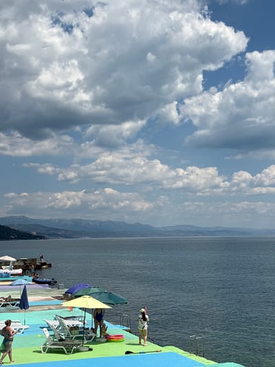 The image depicts a coastal scene with a body of water, distant mountains, and a vibrant, multi-colored platform with beach amenities. The sky is filled with large, fluffy white clouds against a blue background. On the platform, several lounge chairs are set up, some occupied by people relaxing. Beach umbrellas, including yellow, blue, and green ones, provide shade. A few individuals are visible, including a woman in a blue swimsuit standing near a child's inflatable pool, and another woman walking across the platform. Further in the background, a pier extends into the water with people and a small boat. The sea is calm and a muted blue, and the distant mountains appear hazy. The location is specified as Kiparisnoe, Ukraine.