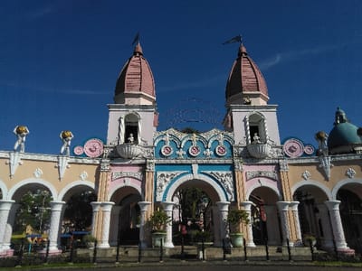 This image depicts an ornate gateway structure located in Batulawang, Indonesia, under a clear blue sky. The multi-tiered, symmetrically designed edifice features two prominent towers capped with conical, light pink domes and dark pennants. The building's facade is primarily composed of light pink, pale yellow, white, and light blue sections, heavily adorned with decorative carvings, scrolls, and circular medallions. A large central archway is flanked by multiple smaller arches, supported by white fluted columns. On the roofline to the left, two white male figures, each bearing a golden spherical object, are visible. Additional smaller white figures are situated within the arched openings of the towers. A teal-colored dome is partially visible behind the structure on the right side. Green foliage is present in the background through the arches, and a low, dark metal fence is in the immediate foreground. No people or specific actions are depicted.