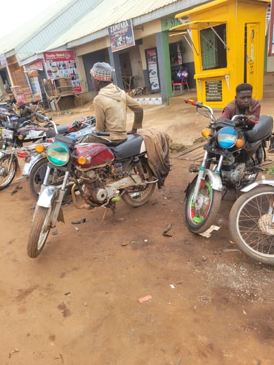 In Bokkos, Nigeria, two individuals are seen with motorcycles. One man, wearing a maroon shirt and brown vest, sits on a motorcycle in front of a bright yellow kiosk. The kiosk features text related to "MTN Products & Services," including SIM registration, data bundles, and phone sales. Another person, dressed in a light brown jacket and a patterned cap, is seated on a different motorcycle parked on the dirt ground. The motorcycles appear to be undergoing some form of repair or are parked for business. The scene suggests an outdoor commercial area, possibly a roadside stall or a small shop. The lighting indicates it is daytime. The ground is dusty and uneven, scattered with debris, typical of an outdoor environment. Signs for "ZAMFARA" and "Emeke Global Multi-purpose Ventures" are visible, indicating commercial activity in the area. The overall impression is one of daily commerce and transportation in a Nigerian town.