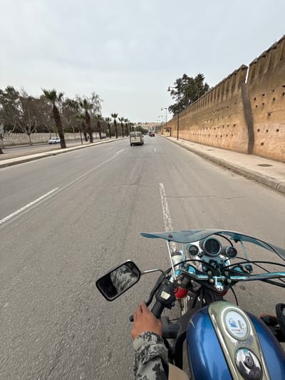 The image depicts a first-person perspective from a motorcycle traveling on a multi-lane asphalt road in Mechouar Fes Jdid, Morocco, under an overcast sky. In the immediate foreground, the blue fuel tank of the motorcycle, a chrome instrument panel with gauges, and a clear windshield are visible. The operator's left hand, clad in a camouflage-patterned sleeve, grips the handlebar and brake lever. A rectangular rearview mirror on the left reflects a distorted view of the rider's upper body.
The road ahead features white dashed and solid lane markings, with visible cracks in the asphalt. Traffic includes a white cargo van directly in front of the motorcycle, and other vehicles (a red car and a dark car) further down the road.
To the right, a wide concrete sidewalk borders a tall, ancient-style fortified wall constructed of tan-colored material, featuring crenellated battlements along its top and multiple small, circular openings/niches at various heights. Several streetlights with dark poles and red-domed fixtures are spaced along the wall.
To the left, a low white wall separates the road from a linear arrangement of palm trees with green fronds and some bare deciduous trees. Beyond these trees, another white wall or building is visible, and a white sedan is present. In the distant background, a cluster of buildings on a slight elevation forms part of the cityscape. The overall environment is urban, suggesting a main thoroughfare.