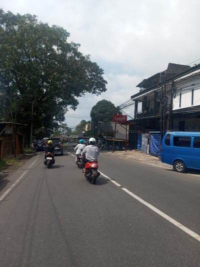 The image depicts a daytime street scene in the city of Niz Lakuk, India, with a clear sky partially obscured by clouds. The asphalt road occupies the foreground and midground, featuring a broken white lane marker on the right side and visible tire streaks on the left. Three individuals on motorcycles are moving away from the camera in the right lane. The closest rider wears a white helmet and light gray long-sleeved shirt, operating a red scooter. Behind them, another rider wears a light blue helmet and white shirt on a dark scooter. A third rider, further back, wears a yellow helmet and dark top on a black scooter. In the background, a black SUV and a blue car are also visible. On the right side of the road, a blue minivan is parked adjacent to a sidewalk area. This area transitions into a building facade partially covered by blue tarps, and further right, a multi-story building with exposed utility wires and poles. A red sign with "LAZATTOH" is mounted on a pole near the buildings. The left side of the road is bordered by dense green foliage from large trees, with some wooden fencing or structures visible beneath them.