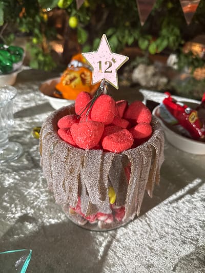 A glass container is filled with red strawberry-shaped marshmallows, edged with brown candy strips covered in sugar. Inside the container, other types of candies are visible. A pink star with the number "12" is attached to the marshmallows. The container sits on a textured white surface near a glass and other objects. In the background, foliage and partially visible party decorations are present.