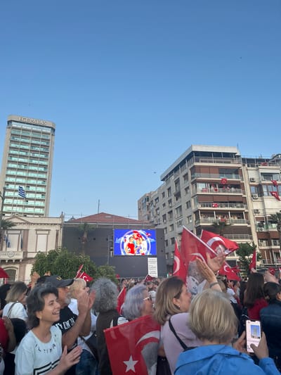A large crowd of people are gathered in the Konak Square of İzmir, Türkiye, cheering and waving Turkish flags. A large screen displays an image of four people, two adults and two children, holding soccer balls. The people in the crowd are mostly young adults and middle-aged adults. Some children are also present. The crowd is standing on the street in front of a large building with a digital billboard. The sky is clear and blue. A tall building with the sign "EGE PALAS" is visible in the background. The image was captured in Konak Square, Izmir, Turkey at 38.437 latitude and 27.141 longitude. The crowd is facing the screen, and some are waving Turkish flags. The people in the crowd are all smiling and laughing, and the atmosphere is festive. Some of the people are wearing hats or scarves, and others are wearing casual clothing. The image is taken from a slightly elevated angle, giving the viewer a good perspective of the crowd and the surrounding buildings. The image is clear and well-lit.