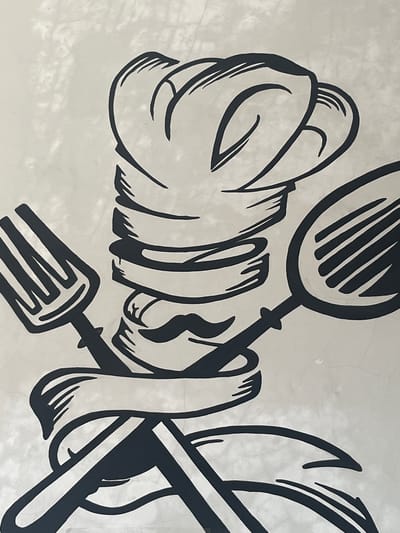 This image features a stylized black and white graphic design, likely a mural or painted artwork, against a textured, light-colored background that appears to be a wall with subtle cracks. The design depicts a hand, rendered in thick black outlines, holding a spoon and a fork that are crossed. The hand is depicted as if it is made of flowing, ribbon-like material, creating a dynamic and abstract representation. The spoon is positioned above and to the left, its bowl facing downwards and featuring parallel lines suggesting texture or shadow. The fork is below and to the right, its tines clearly visible, also with parallel lines. The overall impression is of a culinary theme, conveyed through the prominent kitchen utensils and the abstract rendering of a hand. There are no explicit location cues, time of day, or weather elements visible. The emotions conveyed are abstract due to the stylized nature of the artwork, but the strong lines and dynamic composition could suggest energy or creativity. There is no visible text in the image.