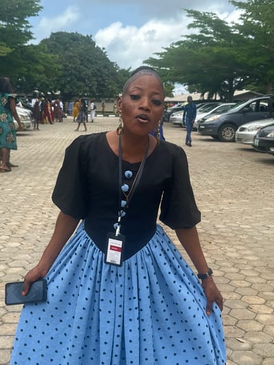 A woman is standing in a parking lot in Itoga, Nigeria. She is wearing a black top with puff sleeves and a light blue skirt with black polka dots. She has on large hoop earrings and a necklace with a pendant. A lanyard with an ID badge is hanging around her neck. She is holding a dark-colored smartphone in her left hand. The parking lot is paved with interlocking stones. In the background, there are several cars parked, and people are visible in the distance, some walking and others standing. Trees line the edge of the parking lot, and the sky is cloudy.