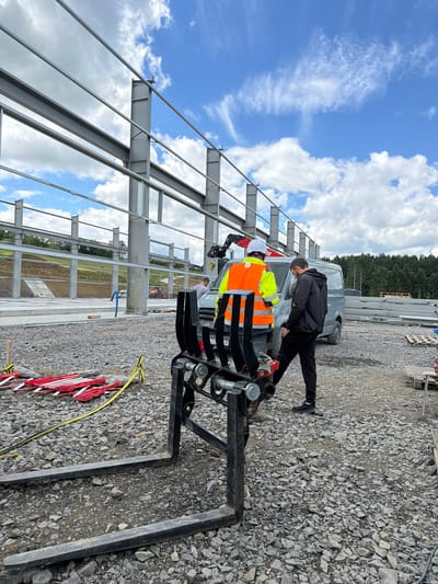 The image depicts an outdoor construction site in Stróża, Poland, with a large steel structure under construction. In the foreground, a forklift attachment with two large prongs rests on a gravel surface. Two men are visible in the midground. One man, wearing a bright orange high-visibility vest and a white hard hat, stands facing away from the camera. He appears to be operating a piece of equipment attached to a van. The second man, dressed in casual dark clothing, stands with his back to the camera, looking towards the first man and the van. A grey van is parked behind the forklift and near the men. In the background, the skeletal framework of a large building is taking shape, supported by concrete foundations and numerous steel beams. Beyond the construction area, a treeline marks the edge of the site, with a gently sloping, grassy hill visible to the left. The sky above is blue with scattered white clouds.