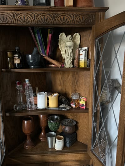 The image shows the inside of a wooden cabinet or cupboard, possibly located in a private residence, filled with various objects. The cabinet has a glass-paneled door at the top. Inside, the shelves contain a diverse collection of items.
On the left shelf, there are decorative angel figures, incense boxes, colored file folders, and a marble mortar and pestle set. At the bottom are two bottles of liquid, likely wine or oil.
The right shelf contains containers with food items, a decorative disc, candle holders, drinking glasses, decorative cups, and a wooden decorative set with brown goblets. The items appear to be a miscellaneous collection of decorative, functional, and household objects.
There is no direct evidence to confirm the location is in Gränsta, Sweden.