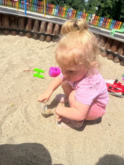 A young blonde girl with her hair in a bun is playing in a sandbox in Donchovci, Bulgaria. She is wearing a pink t-shirt and pink shorts, and is kneeling in the sand. The girl is holding a clear plastic cup and appears to be pouring sand from it. There are several children's toys scattered around the sandbox, including a green "H" shaped toy and a pink toy with holes. In the background, a colorful fence of a playground is visible, along with trees and foliage. The ground is covered in sand, with scattered pebbles and small shells. The overall lighting suggests a sunny day.
