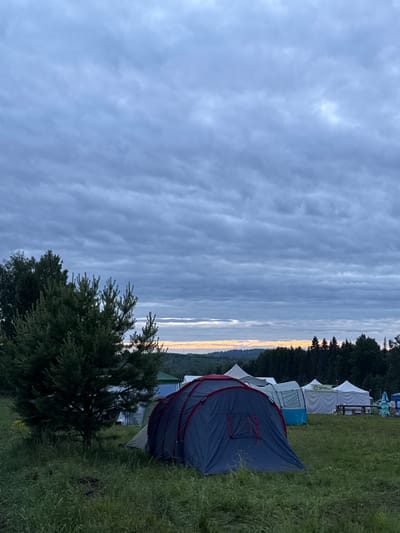 The image depicts an outdoor camping or event site, likely at either dawn or dusk, under an overcast sky. The location is Большая Кивара, Russia.
The upper approximately two-thirds of the frame are dominated by a dense formation of stratocumulus or altocumulus clouds, appearing in shades of blue-grey and dark grey, indicating heavy cloud cover. A narrow horizontal band of light, transitioning from white to orange-yellow, is visible along the horizon line, suggesting the presence of a rising or setting sun beneath the clouds.
The horizon is defined by a dense line of dark green coniferous trees, appearing as a thick forest boundary. Beyond this forest, rolling terrain or distant hills are faintly visible.
The midground and foreground consist of a grassy field. A prominent, dark green coniferous tree, possibly a pine or fir, stands in the left portion of the image, extending from the bottom edge to approximately the mid-frame height.
Multiple tents are set up across the grassy area. In the immediate foreground, a dark blue tent with red structural accents and zippers is centrally placed, facing slightly left. To its right and further back, another blue and white tent is visible. Beyond these, a cluster of white tents, some with pointed roofs (possibly marquees or event tents), are arranged, some appearing connected. A sign with Cyrillic text, reading "КОМНАТА ДЛЯ ЖДАНИЯ" (Room for Waiting), is visible on one of the white tents. Below these white tents, wooden tables and benches are discernible. Two blue, conical figures, possibly garden gnomes or decorative elements, are situated near the white tents on the far right.
The grass in the foreground is varied in length and texture, appearing natural and unmanicured. No individuals are visible anywhere in the scene. The overall lighting is dim and diffused, consistent with twilight or an heavily overcast day.