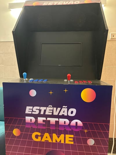 The image displays a custom-built arcade gaming cabinet situated indoors in the city of Aveiro, Portugal. The cabinet's lower section is adorned with a dark purple, retro-themed graphic featuring a pink-purple grid, scattered multi-colored circles, yellow stars, and the prominent text "ESTÊVÃO RETRO GAME" in white, gradient, and yellow fonts respectively. The control panel, black in color, is equipped for two players: a light blue joystick with seven corresponding light blue push buttons on the left, and a red joystick with seven red push buttons on the right. Above the control panel, a black housing contains an Acer flat-screen monitor, with two circular speaker grilles positioned above the screen. A top marquee displays the word "ESTÊVÃO" alongside an orange circular graphic. The immediate environment includes a partial view of a turquoise soft furnishing on the lower left, and light-colored tiled walls with an overhead light fixture visible in the background. No people are present.