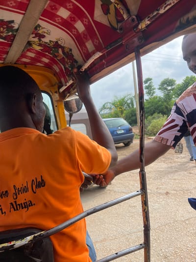 The image shows the interior of a yellow and red motorized rickshaw, a common form of transport in Kuchingoro, Nigeria. The driver, wearing an orange t-shirt with the text "Social Club, i, Abuja" visible, is seated and operating the vehicle. Another person, wearing a patterned shirt, is visible in the foreground, leaning towards the driver. The rickshaw's ceiling is decorated with a red and white checkered pattern and floral motifs. Outside the rickshaw, a blue car is parked on a dirt road, with lush green vegetation and trees in the background under a cloudy sky. The scene depicts everyday life and transportation in a Nigerian city.