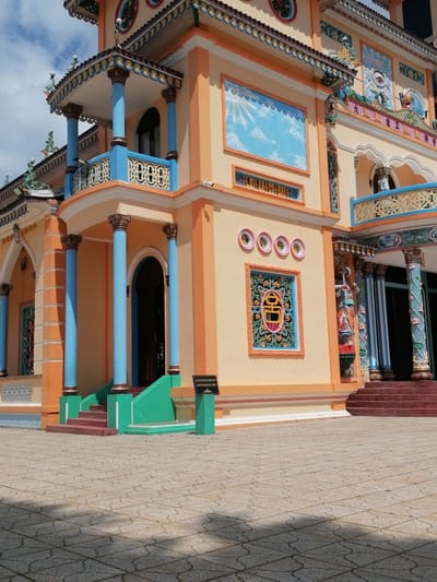 The image shows a colorful building, likely a temple, with orange and yellow exterior walls. The building features ornate decorative elements, including blue and gold trim, blue pillars, and intricate carvings. There are several windows and arched doorways. A set of red stairs leads up to an entrance flanked by blue pillars. The ground in front of the building is paved with square tiles. A black sign with white text is visible near the stairs. The sky above is blue with some white clouds. The image is taken from a low angle, looking up at the building. The context provided indicates the location is in Lộc Quý, Vietnam. No people are visible in the image.