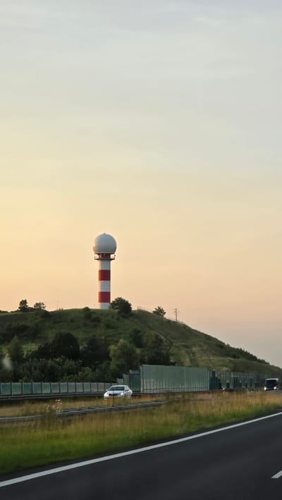 The image shows a Doppler weather radar tower with red and white stripes, topped with a white dome, situated on a grassy hillside. The tower is positioned in the middle ground. To the right of the tower, a highway with white lane markings is visible, running parallel to the hillside. There are several cars on the highway, including a white car in the foreground and another darker car further down the road. A green sound barrier runs along the side of the highway, separating it from the hillside. The hillside is covered in green grass and scattered trees. The sky is a soft gradient of pale yellow and white, suggesting it is either sunrise or sunset. The overall scene appears to be a rural or semi-rural setting in Siemonia, Poland, with the weather radar indicating a focus on meteorological monitoring. There is no visible text in the image. The image is oriented vertically.