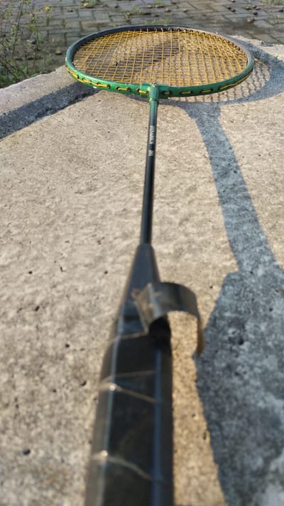 A close-up, low-angle shot shows a badminton racket lying on a textured concrete surface. The racket's head, with its yellow strings and green frame, is visible in the upper portion of the image, casting a shadow to the right. The grip of the racket, wrapped in black tape, extends towards the foreground and is slightly out of focus. The background is blurred, showing hints of dry vegetation and more concrete. The overall lighting suggests it is daytime, with sunlight creating distinct shadows. The scene is set in Trosobo, Indonesia, on a concrete pathway.