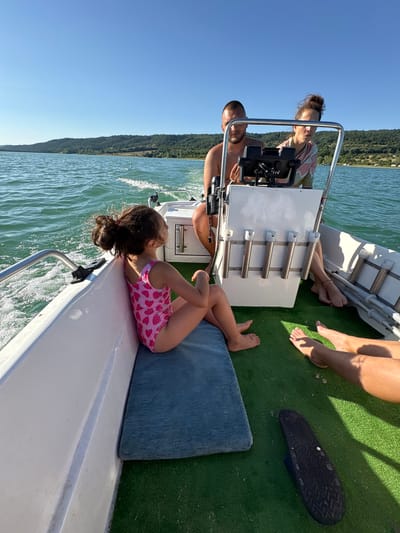 An outdoor, mid-angle, eye-level shot captures a family enjoying a boat ride on a sunny day in the city of Golyama Zhelyazna, Bulgaria. The boat is moving through a body of water with green, choppy waves, and a forested shoreline with hills rises in the background under a clear blue sky. In the foreground, a young girl with dark hair pulled back in a ponytail sits facing the stern of the boat, wearing a pink swimsuit adorned with strawberries. She is seated on a dark blue cushion on the green carpeted floor of the boat. To her right, two adults, a man and a woman, are seated towards the front of the boat, facing forward. The man, with short hair and a beard, is partially obscured by the woman. The woman, with her hair tied up, is looking towards the boat's console, which has a Garmin GPS device. Their legs are visible, and the woman is wearing a light-colored, patterned top. Other individuals' legs are also visible in the foreground on the right, indicating at least two more people are on the boat. A black sandal lies on the green carpet near the right edge of the frame.