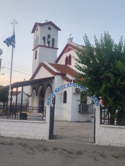 The image shows a white church with red trim in Νέα Ραιδεστός, Greece. A Greek flag flies from a flagpole to the left of the church, with a cross on top. The church has a bell tower with arched openings and a red-tiled roof. An arched sign in blue letters above the entrance reads "ΙΕΡΟΣ ΝΑΟΣ ΑΓΙΟΥ ΘΕΟΔΩΡΟΥ". The church is surrounded by a low white brick wall and a black metal fence. A tree with green leaves is to the right of the entrance. The sky is clear and pale blue, suggesting it is either early morning or late afternoon. An air conditioning unit is mounted on the exterior wall of the church. The ground in front of the church is paved with grey tiles and asphalt.