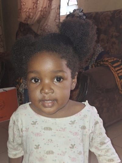 A young child, approximately 1-3 years old, with dark skin and dark, Afro-textured hair styled into two prominent puffs, faces forward. The child wears a light-colored, long-sleeved top patterned with small pink elephants and other indistinct shapes. The child's gaze is directed towards the viewer with a neutral expression and slightly parted lips. The setting is an indoor residential space in Kaduna South, Nigeria. In the background to the right, a portion of a brown upholstered sofa or armchair is visible, draped with a patterned cloth featuring black, orange, and yellow geometric designs. A translucent brown curtain with a floral pattern partially covers a window in the upper right background. To the left, a segment of an orange bag or box with white lettering is visible. The floor is covered with light-colored tiles. Ambient lighting illuminates the scene.