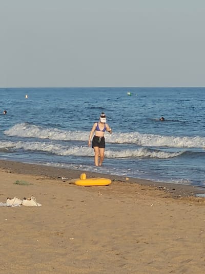 A woman wearing a blue bikini top, black shorts, and a white visor walks in the shallow water near the shore of a beach. She is holding a white object in her right hand. Two sets of white sneakers are on the sand in the foreground, to the left. A yellow inflatable ring is partially visible on the sand to the right of the woman. The sea is blue with white waves, and the sky is clear and pale blue. Two buoys are visible in the water, one yellow and one green and white. Another person is partially submerged in the water further out in the sea. The scene appears to be at Platja de Castelldefels, Spain.