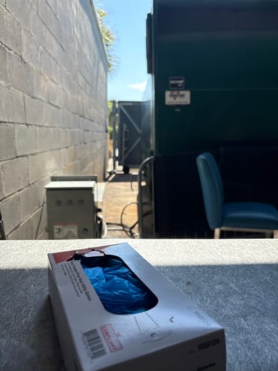 In Panama City Beach, Florida, an outdoor scene is depicted with a focus on a box of blue disposable gloves in the foreground, placed on a speckled, light-colored surface. Behind the gloves, a narrow alleyway is visible, bordered by a grey brick wall on the left and a large dark green dumpster on the right. The dumpster has two small rectangular labels on its side, one white with red lettering and another black with white text. Further down the alley, a dark trailer or truck bed is partially visible, and beyond that, a light blue chair with a metal frame sits near the dumpster. Palm fronds extend from behind the brick wall into the bright blue sky, suggesting a sunny day. The overall impression is that of a back service area, possibly behind a commercial establishment.