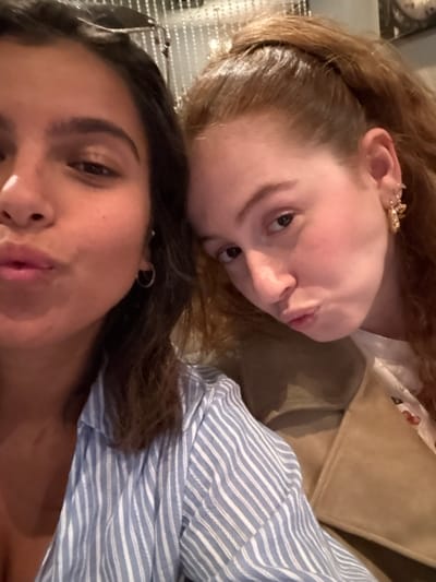 The image depicts two adult females posing for a close-up selfie indoors. The specific location within the city of West Windsor, United States, cannot be determined from the visual evidence. The female on the left has dark brown hair and is wearing a light blue and white striped shirt with small silver hoop earrings. She looks directly at the camera with lips pursed. The female on the right has reddish-brown hair styled in a high ponytail and is wearing a light-colored top, possibly with a subtle cherry pattern, beneath a tan jacket. Her right ear features multiple gold-colored earrings, including a hoop and a decorative charm. She is positioned slightly to the right, looking towards the left, also with pursed lips. The background is out of focus, showing a shimmering, possibly beaded or metallic, decorative surface behind the left female, and a darker, indistinct area to the right. The scene is illuminated by what appears to be warm, artificial lighting.