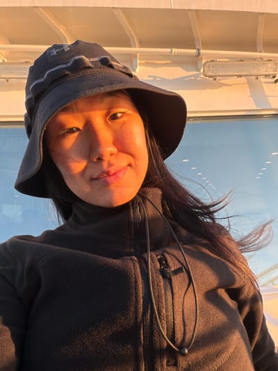 A woman, wearing a dark bucket hat and a dark zip-up jacket, is pictured close-up in an outdoor setting. The sun is shining, casting a warm, golden light on her face, particularly on the right side of her face, and on her jacket. Her hair is dark and appears windblown, with strands of it across her face and blowing away from her head. In the background, there's a large window or glass surface reflecting the blue sky with some oval-shaped lights. Above the window, there's a white structure with horizontal pipes and a light fixture, suggesting the location might be a boat or a building with a similar architectural style. The woman is looking towards the camera with a slight smile. The overall impression is that of a casual, sunlit portrait taken outdoors, possibly on a ferry or a waterside structure.