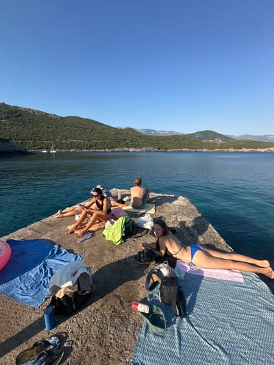 The image depicts a group of people relaxing on a concrete pier by the sea. The setting is a picturesque coastal area with green, tree-covered mountains in the background and clear blue water. The sun is high in the sky, suggesting it's midday or early afternoon, and the weather appears to be warm and sunny.
Several individuals are visible, mostly lying down on towels or directly on the pier, basking in the sun. One person is lying on a blue and white patterned blanket, wearing a blue bikini. Another person is seated, wearing a black bikini. There are various objects scattered around, including towels of different colors (blue, green, pink), bags, sandals, and a can of what appears to be a beverage. A pink inflatable ring is also visible near the edge of the pier. The overall mood seems relaxed and leisurely, with people enjoying a day at the beach or by the water.
The location is identified as Dubraljevina, Montenegro. The scene is characterized by the contrast between the rugged concrete pier, the calm blue sea, and the lush green mountains. The lighting indicates a bright, sunny day.