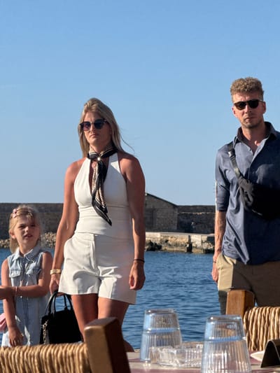 The image depicts three individuals standing outdoors near a body of water, likely the sea, with a stone structure in the background. The setting appears to be a waterfront area in Chania, Greece, possibly a promenade or a restaurant area.
In the foreground, a woman with blonde hair is walking, wearing a white halter top, white shorts, and sunglasses. She has a patterned scarf around her neck and is holding a black handbag. To her left, a young girl with blonde hair and a pink clip is wearing a denim dress and sunglasses. To the right of the woman, a man with curly blonde hair is standing, wearing a blue collared shirt, sunglasses, and khaki shorts. He has a black fanny pack slung across his chest.
In the middle ground, the water is visible, and a stone wall or embankment lines the shore. The background shows more of the stone structure and a clear blue sky. The lighting suggests it is daytime, with the sun casting shadows.
On a table in the lower foreground, two empty drinking glasses are placed on a surface covered with what appears to be wicker or rattan material. A small ashtray is also visible. The overall scene conveys a casual, daytime outing in a scenic coastal location.