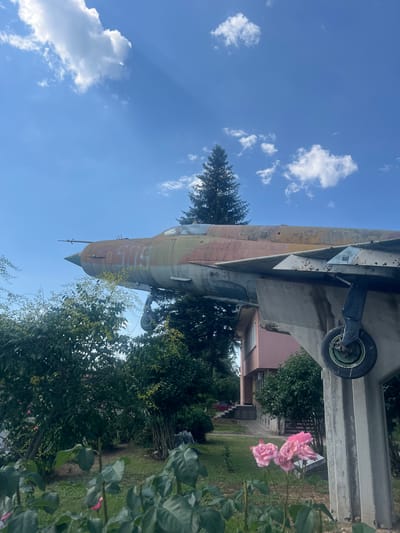 An old, weathered jet fighter, painted in camouflage colors and bearing the markings "509," is mounted on a concrete pedestal in Zelenikovo, Bulgaria. The aircraft is positioned outdoors, amidst lush green trees and bushes. In the foreground, several pink roses are in bloom. A light-colored building with a pink facade and dark windows stands in the background, partially obscured by the trees and the jet. The sky above is a vibrant blue, dotted with fluffy white clouds, with sunlight casting rays through the gaps. The overall scene suggests a memorial or a display of historical military hardware in a public or semi-public space.