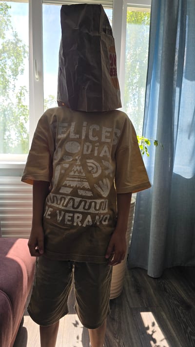 An individual, appearing to be a child, stands centrally in a well-lit indoor space within the city of Oktyabr'skii, Russia. The person's face is completely obscured by a crumpled brown paper bag, likely from a Burger King restaurant, with remnants of the fast-food chain's logo and branding visible on its surface. The individual wears a light tan or yellowish-brown short-sleeved t-shirt with distressed white text and graphics, reading "FELICES DÍA" (Happy Day) at the top and "DE VERANO" (of Summer) at the bottom, interspersed with abstract tribal-style patterns, including a stylized teepee or mountain, circular motifs, and handprint-like shapes. Below the t-shirt, the person wears dark olive green or brown shorts. The hands are visible at the sides, hanging naturally.
The background features a large window on the left, admitting significant natural light, which highlights green foliage and parts of trees outside. A clear sky is visible through the upper portion of the window. On the right side of the frame, a translucent light blue or grey-blue curtain is partially drawn, partially obscuring a view that includes more greenery and a light-colored, possibly woven, cylindrical object on the floor. A portion of a pink or mauve upholstered armchair or sofa is visible in the bottom-left corner. The floor is covered with dark wood-laminate or engineered wood, onto which distinct shadows are cast from the window frames and what appear to be leaves, indicating strong direct sunlight.