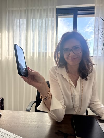 A woman wearing glasses and a white blouse is holding a smartphone. She is wearing gold bracelets and a gold necklace. She is seated at a desk with a notebook and keyboard visible. A window with blinds is behind her. The location in Torres Vedras, Portugal cannot be determined from the image.