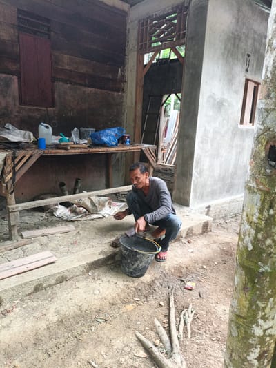 A man is crouched down in front of a partially constructed building in Meunasahuket, Indonesia. He is wearing a grey long-sleeved shirt and dark pants, with red sandals on his feet. In front of him is a black bucket. He is holding a trowel-like tool in his right hand and appears to be scraping something from the edge of the bucket. The ground is dirt with some scattered debris and a few pieces of wood. Behind him is a wooden workbench covered with various items, including plastic containers, bags, and tools. The building behind him is made of rough concrete and wood, with an open doorway revealing the interior. A tree trunk is visible on the right side of the frame. The scene suggests construction or repair work is in progress.