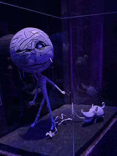 The image shows a display case containing two mummy-like figures, illuminated by purple lighting. The larger figure, on the left, is standing and appears to be a stylized humanoid with a large, round head wrapped in bandages, a wide grin, and a single visible eye. Its body is thin and also wrapped in bandages, with spindly limbs. A smaller, creature-like figure, resembling a stylized armadillo or rhino with a long snout, is positioned on the right side of the display, also wrapped in bandages. The figures are on a dark, textured surface, and the background is dark and indistinct, suggesting an indoor environment, possibly a museum or an exhibition. There is a reflection visible in the glass of the display case, showing a green, one-eyed creature, likely another character from the same fictional universe. The setting is not identifiable as being in Monterrey, Mexico.