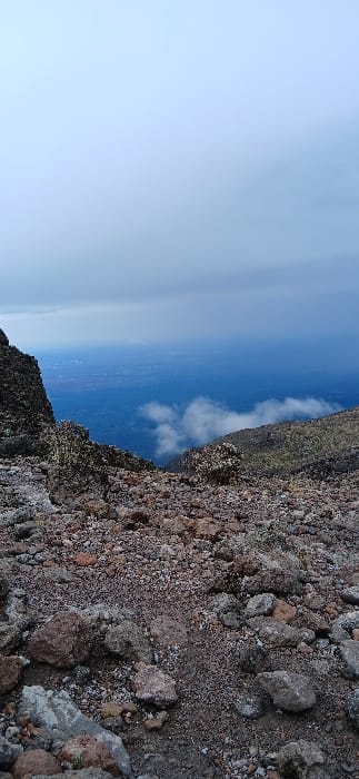The image depicts a high-altitude, rugged mountain landscape at Gilman's Point, Tanzania. The foreground is dominated by a rocky, scree-covered slope composed of various sizes of volcanic rocks in shades of grey, brown, and reddish-orange. A narrow, dirt path is faintly visible winding through the loose rock and gravel. Sparse, low-lying, hardy vegetation, possibly lichens or stunted shrubs, clings to some rock formations and patches of ground.
Below the immediate foreground, the terrain drops steeply into a vast expanse. Clouds are visible below the viewer's elevation, partially obscuring the midground. The distant background reveals a hazy, expansive view of what appears to be plains or lower elevation land, tinted blue by atmospheric perspective. Faint patterns consistent with distant human settlement or urban areas can be discerned on the horizon line in the far distance. The sky above is heavily overcast with a uniform grey-blue cloud cover, indicating diffused light conditions. No people, animals, or artificial structures are present in the immediate or mid-ground.