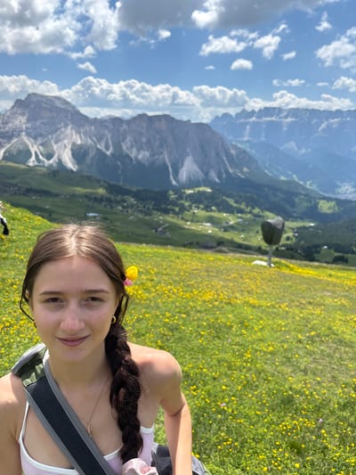 A young woman with brown hair braided into a plait, and a yellow flower tucked behind her ear, is in the foreground of this image. She is looking towards the camera with a slight smile. She is wearing a light-colored top and a backpack. Behind her, a vast meadow covered in yellow wildflowers slopes down into a valley. In the background, majestic mountains rise under a bright blue sky dotted with white clouds. The scene is identifiable as Santa Cristina Gherdëina - St. Christina in Gröden - Santa Cristina Valgardena, Italy, a location known for its stunning alpine scenery and meadows.