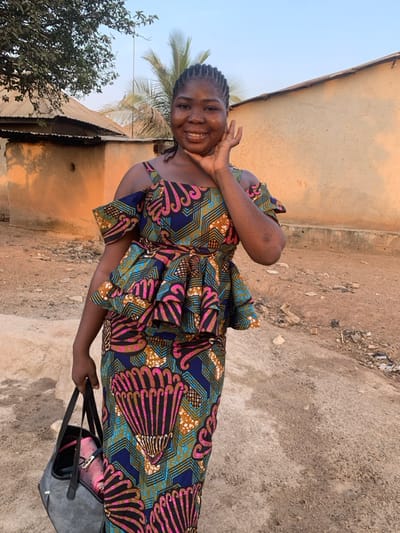 A smiling woman in a vibrant, patterned dress poses outside. The dress features a mix of navy, teal, brown, and pink designs, cinched with a matching sash. She holds a bag, and a small house with a corrugated metal roof is partially visible in the background, along with some vegetation. The terrain appears to be dirt and dust. The sky is bright and clear.