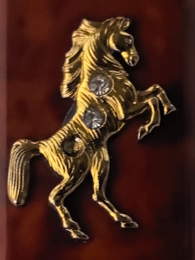 The image shows a close-up, angled view of a golden horse figurine against a dark reddish-brown background. The horse is depicted in a rearing pose, with its front legs raised. It has a textured mane and tail, and appears to be adorned with three circular, clear stones embedded in its body. The lighting highlights the metallic sheen of the figurine and creates reflections. There are no people or discernible environmental features beyond the background. The location in Ново-Талицы, Russia is not depicted or suggested by the image content.