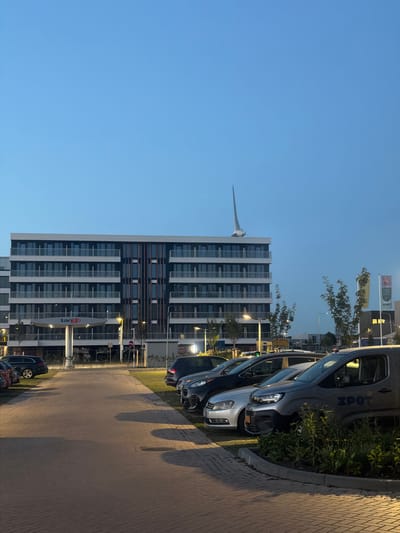 The image captures a dusk scene in Maasdijk, Netherlands. A modern, multi-story building with a contemporary facade featuring numerous windows and balconies dominates the background. A distinctive, white, fin-like sculpture is positioned on the rooftop of the building, extending upwards into the clear blue sky. In front of the building, a Tango gas station canopy is visible. A row of parked cars, including a silver sedan, a dark SUV, and a grey van with "ZPOT" text on its side, are lined up in a paved parking lot. The ground is paved with interlocking bricks, and scattered vegetation and streetlights add to the urban setting. The sky is a gradient of light blue, indicating the transition from day to night.