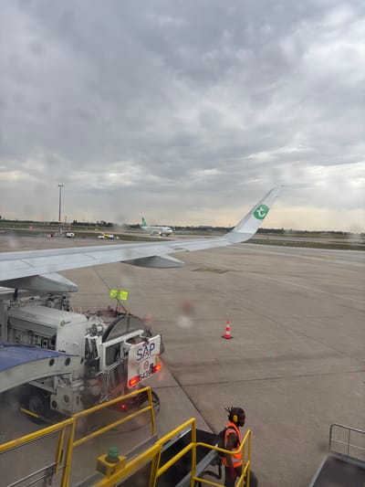 This is a forensic, matter-of-fact description of a scene at Paris-Orly Airport in Paray-Vieille-Poste, France.

The image captures a view from an aircraft window, looking out onto the tarmac on an overcast day. In the foreground, the wing of the aircraft the photographer is in is visible, extending from the left side of the frame and curving towards the right. Adjacent to the aircraft's wing is an airport service vehicle, likely a de-icing or refueling truck, parked on the tarmac. The vehicle is white with blue accents and has various hoses and equipment attached. A worker in an orange high-visibility vest and headphones is visible on the stairs leading down from the aircraft, facing to the right.

Further out on the tarmac, another aircraft with the "Transavia" logo on its tail is taxiing. Several other vehicles, including smaller trucks and possibly airport equipment, are present on the airfield. In the background, airport infrastructure, including lights and buildings, are visible against a sky filled with heavy, gray clouds. The ground is wet, suggesting recent rain or ongoing precipitation. The overall scene depicts a typical operational moment at a busy international airport.