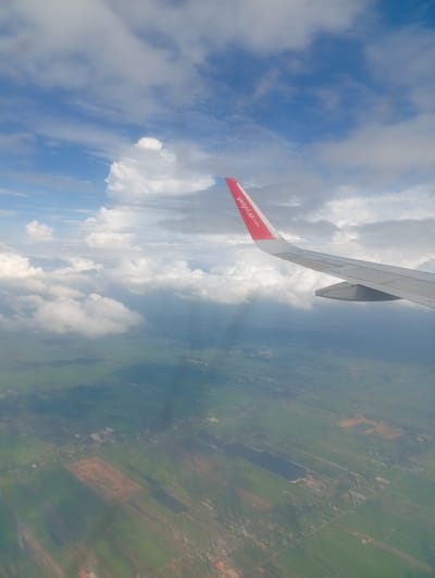 An aerial view captures an airplane wing, emblazoned with the Vietjet Air logo, slicing through a cloudy sky. Below, a patchwork of green fields and darker bodies of water unfolds, indicative of agricultural land. The sun's rays pierce through the clouds, casting ethereal beams onto the landscape. While the specific location within Nong Prue Subdistrict, Thailand, cannot be determined from the image alone, the scene broadly depicts rural scenery as viewed from an aircraft.