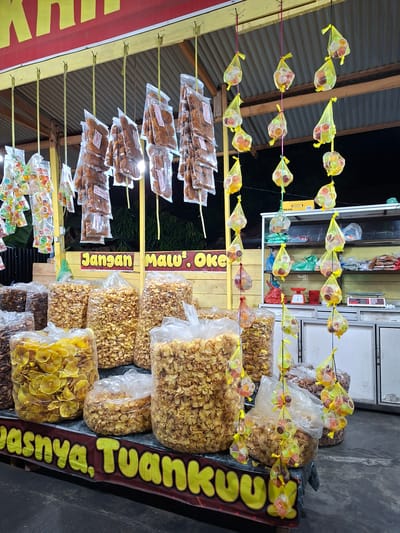 This image captures a nighttime food stall in Glee Gogo, Indonesia, displaying various snacks for sale. The scene is illuminated by artificial lighting, indicating operation after dark.
In the lower foreground, multiple large, clear plastic bags are filled with deep-fried chips; prominent among these are golden-yellow, thinly sliced circular chips, likely banana or cassava, alongside bags containing darker, irregularly shaped chips. A red banner with yellow, block-lettered text, partially visible at the bottom, reads "...asinya. Tuankuu."
Above these large bags, a yellow wooden beam supports numerous small, clear plastic bags containing reddish-brown, crispy savory snacks, possibly fried noodles or similar items, suspended by thin yellow ribbons. To the far left, additional small clear plastic bags containing assorted colorful jelly candies are also hung.
Centrally positioned, a yellow wooden structure frames a red banner with yellow, capitalized text stating "Jangan Malu'. OKE" (Don't be shy. OK).
To the right, a vertical arrangement of colorful jelly candies is displayed. Each candy, encased in a small clear cup, is individually held within a yellow mesh net and suspended by yellow and pink strings at various heights. Behind these hanging sweets, a white, multi-shelf cabinet is visible. The shelves hold various items, including plastic bags, a red mechanical weighing scale, and a modern digital scale. A yellow box marked "TBG-516" rests on top of the cabinet.
The background is predominantly dark, with hints of green foliage beyond the stall. The stall's roof is constructed from corrugated metal. No individuals are visible within the frame.