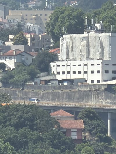 This aerial view captures a dense urban and natural landscape in Gondomar, Portugal, likely during daylight with clear weather. The scene is dominated by a cluster of buildings, some appearing under construction or renovation due to scaffolding and white tarps covering large sections. A tall, multi-story white building with numerous dark, rectangular windows stands prominently.
Adjacent to the buildings is a long, grey concrete structure that appears to be a raised walkway or elevated platform, possibly a viaduct or embankment. A thin, dark line running along its length suggests tracks or a similar linear feature. A small, white vehicle with a blue stripe is visible on this elevated structure.
Further into the scene, traditional Portuguese architecture is evident with buildings featuring red-tiled roofs. Dense green foliage, indicative of trees and vegetation, is interspersed throughout the urban area, suggesting a well-established park or residential gardens. The overall impression is one of a developed area with a mix of modern and older structures, partially integrated with natural elements. No people are clearly discernible, and no text is visible in the image.
