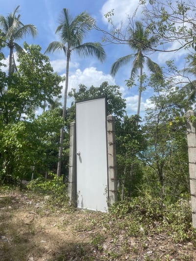 The image depicts a white, double-leafed door standing upright, unsupported, amidst lush tropical vegetation and palm trees. The door is positioned between two concrete posts that appear to be part of a fence, with barbed wire strung between them. The ground in front of the door is dry and covered with sparse vegetation and debris. In the background, dense foliage, palm trees, and a bright blue sky with scattered clouds are visible. The scene suggests an outdoor setting in a tropical climate. The location is identified as Baan Harn, Thailand.