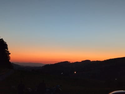 At dusk, a panoramic view captures rolling hills under a gradient sky transitioning from soft blue to vibrant orange. Silhouetted against the horizon are the rounded shapes of trees and the jagged outlines of a forest. Two figures are seated in the foreground, their forms dark and indistinct. A motorcycle is parked nearby. In the middle ground, a distant light glows, possibly from a settlement. The scene appears to be from a vantage point overlooking a valley, consistent with the hilly terrain found in St. Peter, Germany.