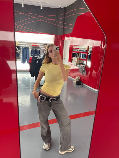A light-skinned female takes a mirror selfie inside a retail clothing store in Abu Dhabi, United Arab Emirates. She is centrally positioned, wearing a fitted light yellow short-sleeved t-shirt, loose-fitting faded gray jeans, a black belt with two large silver oval buckles, and silver-toned athletic sneakers with white soles. Her left hand rests on her hip, revealing a thin silver bracelet, while her right hand holds a light-colored smartphone to capture her reflection. A silver-colored ring is visible on her right ring finger.
The reflected store environment features a gray floor with a thin red stripe. Walls are composed of glossy red panels and matte dark gray sections decorated with thin, wavy pink-red lines. Overhead, white track lighting fixtures and a small black security camera are visible. In the background, clothing racks display various garments, including blue jeans and dark shirts. A white mannequin torso wearing blue jeans is seen on the left. On the right, shelves contain folded clothing and several fabric shopping bags. A male individual, partially visible, stands in the mid-ground, wearing a dark top and holding a magazine or catalog.