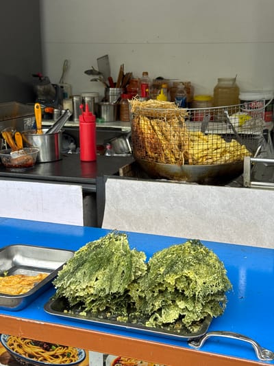 The image shows a food preparation area, likely in a restaurant or food stall. There are various cooking implements, including a deep fryer with food inside a wire basket, containers of sauces, and other kitchen utensils. A tray of what appears to be fried seaweed or a similar crispy, green food item is on the right side of the image. Below the seaweed is a tray with fried food. The overall scene is utilitarian and focused on food preparation. There is no indication of the location in Zhuoshui, China.