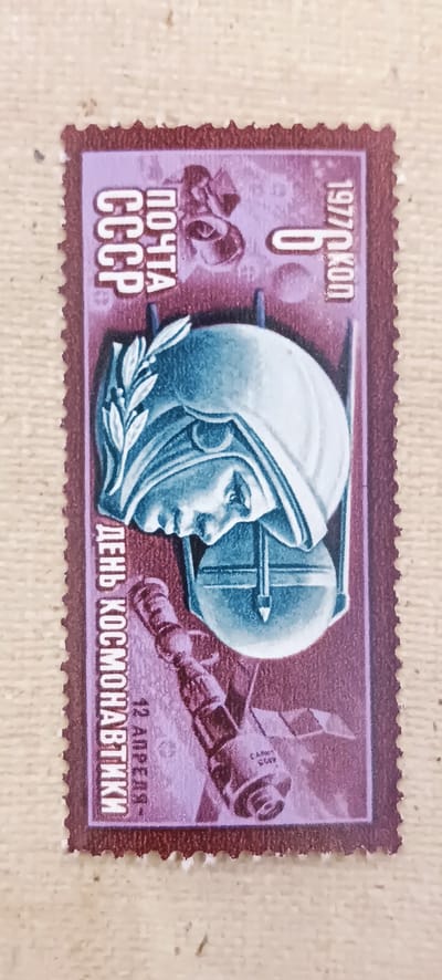 The image is a close-up of a postage stamp with a dark purple border and a lighter purple background. The stamp features a stylized depiction of a helmeted head in profile, facing left. A laurel wreath adorns the helmet. To the left of the helmet, in vertical text, are the words "ПОЧТА" and "СССР" in white capital letters. To the right of the helmet, in vertical text, are "1977/10КОП". Below the helmet and to the left, also in vertical text, are the words "ДЕНЬ КОСМОНАВТИКИ" and "12 АПРЕЛЯ-". In the lower right quadrant of the stamp, a rocket or satellite-like object is depicted. The overall design is commemorative, likely celebrating Cosmonautics Day. There is no information or visual element within the image that pertains to the city of Onaldy, Turkmenistan.